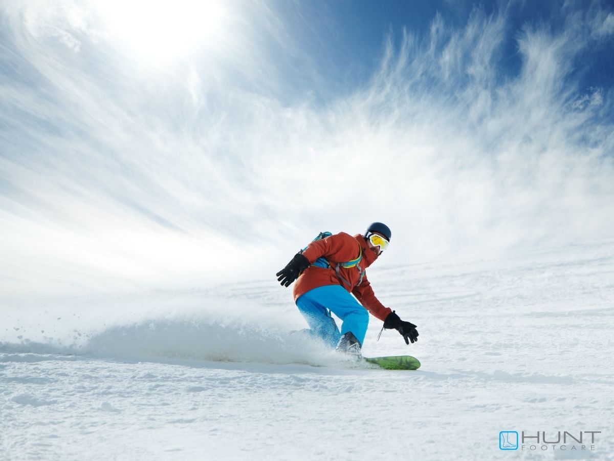 A snowboarder wearing a red jacket, blue pants, and yellow goggles glides down a snowy slope, kicking up powder under a bright, partly cloudy sky—reminding us of the importance of care and prep during the spring snow. Advice from Hunt Footcare logo in bottom right.